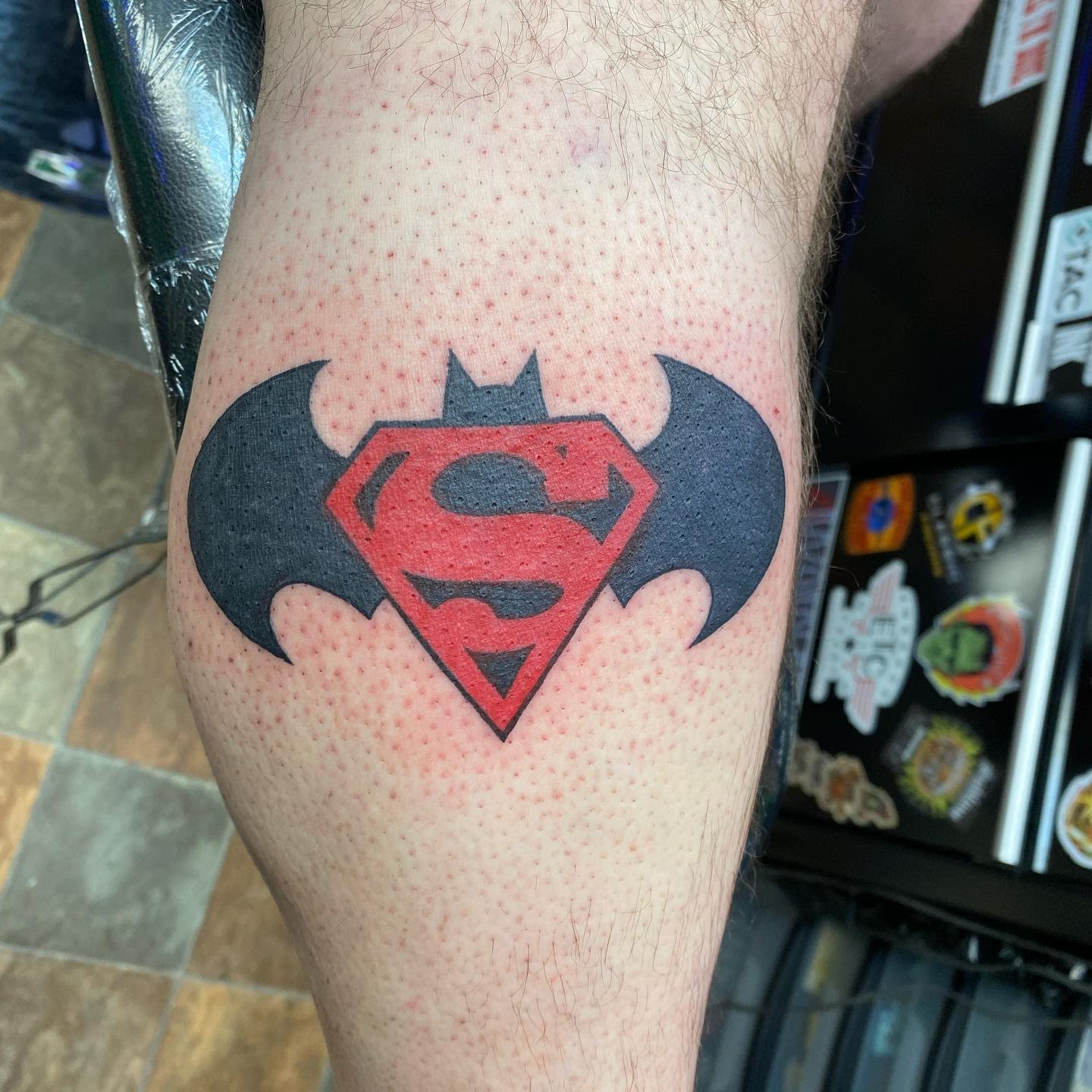Tattoo of Superman and Batman symbols combined on a calf